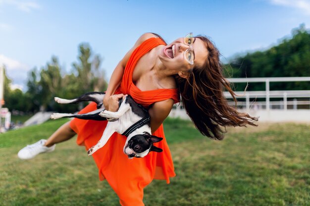 Glücklicher hübscher Frauenpark, der Boston-Terrier-Hund hält, positive Stimmung lächelnd, trendiger Sommerstil, orange Kleid, Sonnenbrille tragend, mit Haustier spielend, Spaß habend, bunt, langes Haar winkend