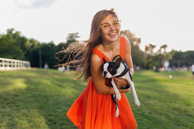 Glücklicher hübscher Frauenpark, der Boston Terrier-Hund hält, positive Stimmung lächelnd, trendiger Sommerstil, orange Kleid, Sonnenbrille tragend, spielt mit Haustier, Spaß, sonnige Wochenendunterhaltung hat