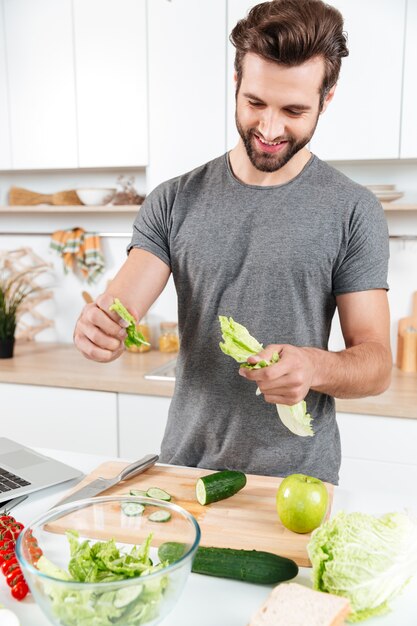 Glücklicher hübscher Kerl, der Salat mit Laptop kocht