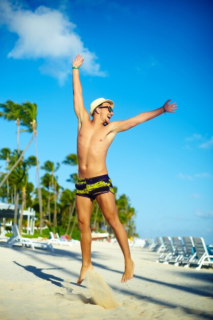 Glücklicher hübscher Mann mit Muskeln im sunhat auf dem Strand, der hinter blauen Himmel hinter blauen Himmel springt