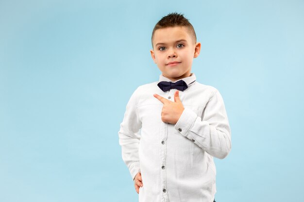 Glücklicher jugendlich Junge, der lokalisiert auf blauem Studiohintergrund lächelt. Schönes männliches halblanges Porträt. Junges emotionales Kind, das nach links zeigt. Das