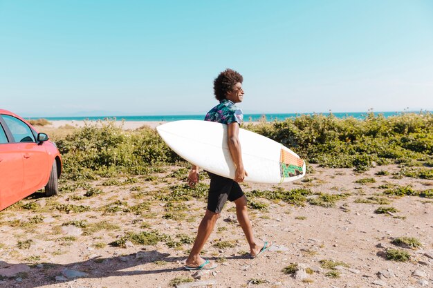 Glücklicher junger Afroamerikanermann, der mit Surfbrett entlang Küste kommt