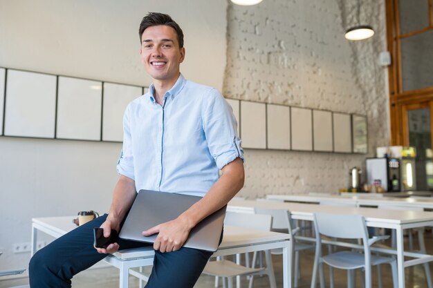 Glücklicher junger attraktiver lächelnder Mann, der im zusammenarbeitenden offenen Büro sitzt und Laptop hält,