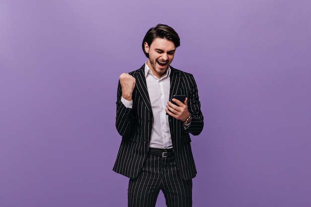 Glücklicher junger brünetter Mann in weißem Hemd und schwarz gestreiftem Anzug, der emotional auf das Telefon schaut, sich freut und gegen die violette Pastellwand posiert