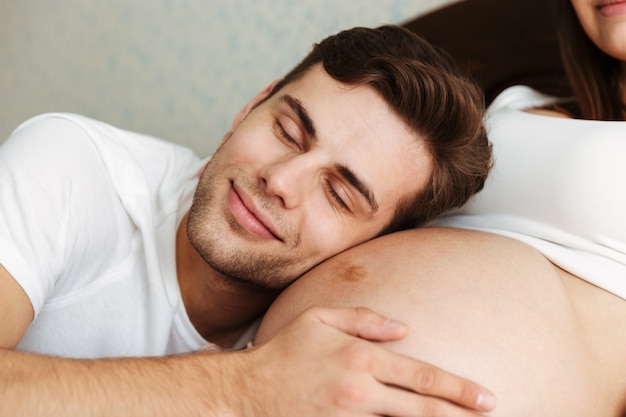 Glücklicher junger Ehemann, der auf dem Bauch seiner schwangeren Frau liegt
