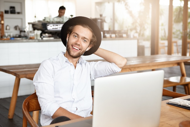 Glücklicher junger Hipster, der weißes Hemd und stilvollen Hut trägt, der fröhlichen Gesichtsausdruck hat