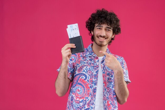 Glücklicher junger hübscher lockiger reisender Mann, der Flugtickets und Geldbörse zeigt und auf sie auf lokalisiertem rosa Raum mit Kopienraum zeigt