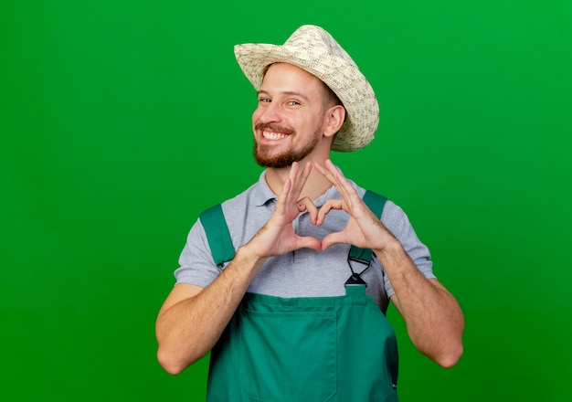 Glücklicher junger hübscher slawischer Gärtner in der Uniform und im Hut, der Herzzeichen tut
