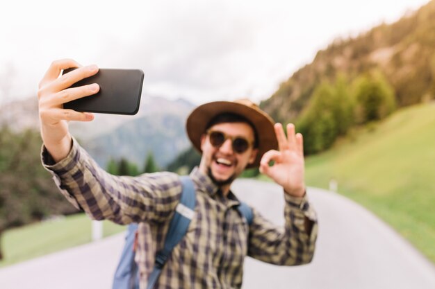 Glücklicher junger Mann, der blauen Rucksack trägt, der selfie mit Lächeln und in Ordnung Zeichen macht, auf der Straße stehend