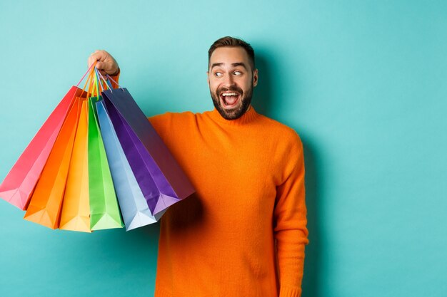 Glücklicher junger Mann, der einkaufen geht, Taschen hält und aufgeregt schaut, im orangefarbenen Pullover stehend gegen türkisfarbene Wand