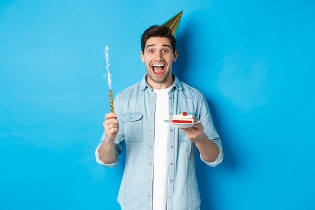 Glücklicher junger Mann, der Geburtstag im Partyhut feiert, B-Day-Kuchen hält und lächelt, auf blauem Hintergrund stehend