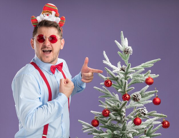 Glücklicher junger Mann, der Hosenträgerfliege im Rand mit Weihnachtsmann und roten Gläsern trägt, die neben Weihnachtsbaum stehen, der mit Zeigefinger darauf zeigt und Kamera betrachtet, die über lila Hintergrund lächelt