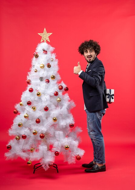 Glücklicher junger Mann, der sein Geschenk hinter dem Stehen hält, das ok Geste nahe geschmücktem weißem Weihnachtsbaum auf der rechten Seite von Rot macht