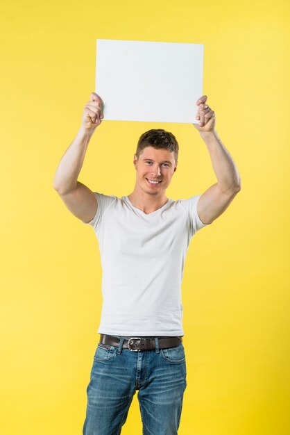 Glücklicher junger Mann, der seine Arme zeigt weißes Plakat gegen gelben Hintergrund anhebt