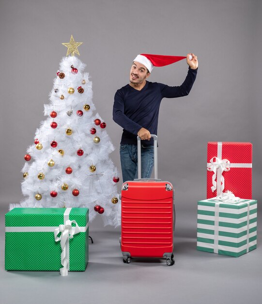 glücklicher junger Mann, der seine Weihnachtsmütze hält, die nahe weißem Weihnachtsbaum auf grau steht