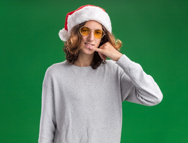 Glücklicher junger Mann, der Weihnachtsweihnachtsmütze und gelbe Brille trägt, die Kamera macht, rufen mich Geste, die über grünem Hintergrund steht