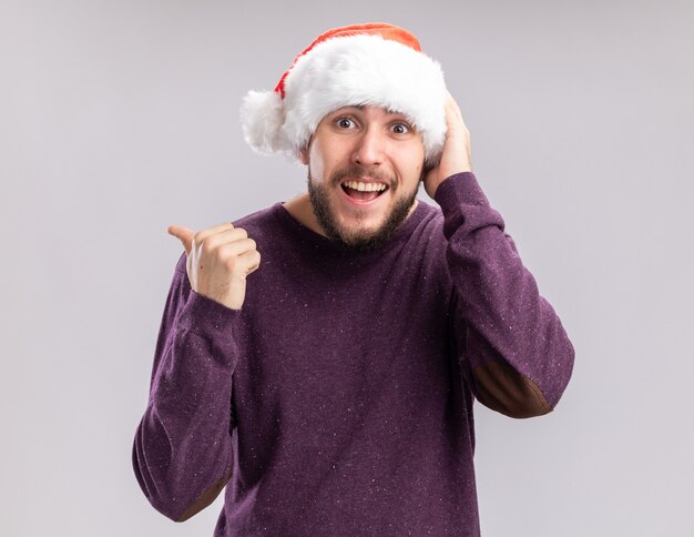 Glücklicher junger Mann im lila Pullover und in der Weihnachtsmannmütze, die Kamera betrachten, die fröhlich zeigt, mit Daumen zurück stehend über weißem Hintergrund