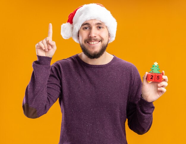 Glücklicher junger Mann im lila Pullover und in der Weihnachtsmannmütze, die Spielzeugwürfel mit Nummer fünfundzwanzig lächelnd zeigt, der fröhlich Zeigefinger zeigt, der über orange Wand steht