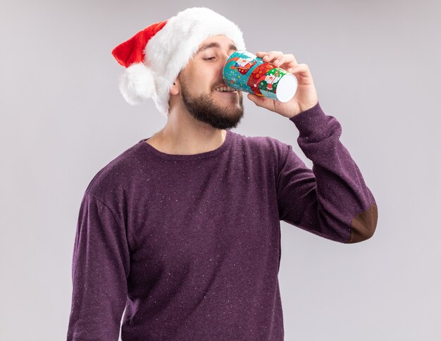 Glücklicher junger Mann im lila Pullover und in der Weihnachtsmannmütze, die vom bunten Pappbecher trinken, der über weißem Hintergrund steht