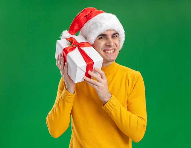 Glücklicher junger mann in gelbem rollkragenpullover und weihnachtsmütze mit lustiger krawatte, die ein geschenk hält, das kamera betrachtet, die fröhlich über grünem hintergrund steht