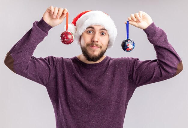 Glücklicher junger Mann in lila Pullover und Weihnachtsmütze mit lustiger Brille, die Weihnachtskugeln hält und mit einem Lächeln auf dem Gesicht auf weißem Hintergrund in die Kamera schaut