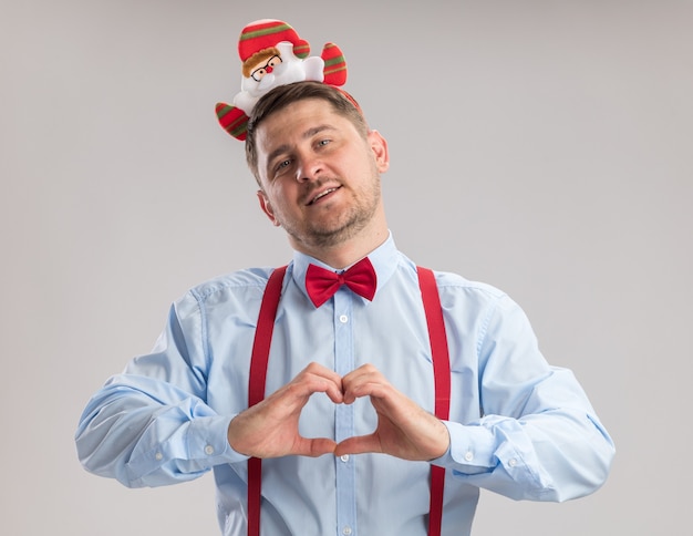 Kostenloses Foto glücklicher junger mann mit hosenträgerfliege in rand mit weihnachtsmann, der die kamera anschaut und herzgeste mit den fingern macht, die fröhlich über weißem hintergrund lächeln