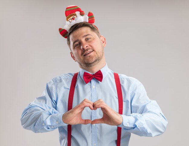 Glücklicher junger Mann mit Hosenträgerfliege in Rand mit Weihnachtsmann, der die Kamera anschaut und Herzgeste mit den Fingern macht, die fröhlich über weißem Hintergrund lächeln