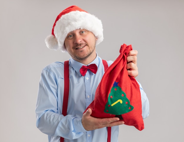 Glücklicher junger Mann mit Hosenträgerfliege in Weihnachtsmütze, der eine rote Weihnachtsmann-Tasche voller Geschenke zeigt und in die Kamera schaut, die über weißem Hintergrund lächelt