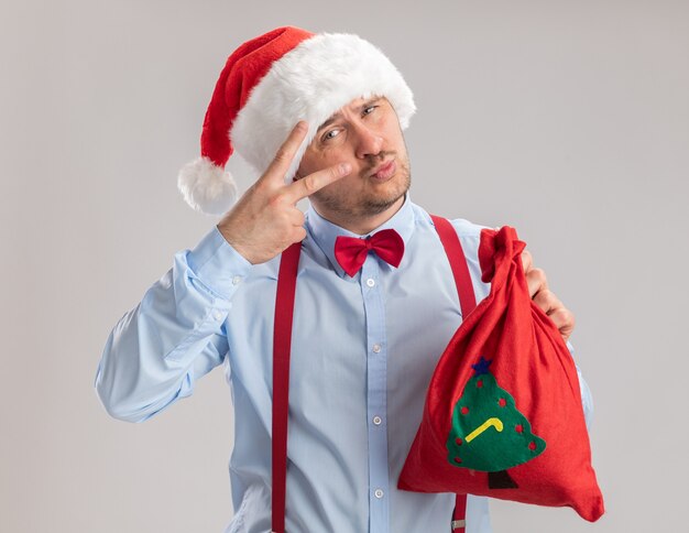 Glücklicher junger Mann mit Hosenträgerfliege in Weihnachtsmütze mit Weihnachtsmann-Tasche voller Geschenke mit Blick in die Kamera mit V-Zeichen auf weißem Hintergrund over
