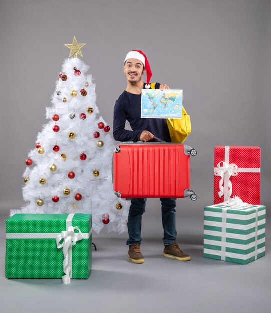 glücklicher junger Mann mit Rucksack, der Karte nahe Weihnachtsbaum hält und auf Grau präsentiert