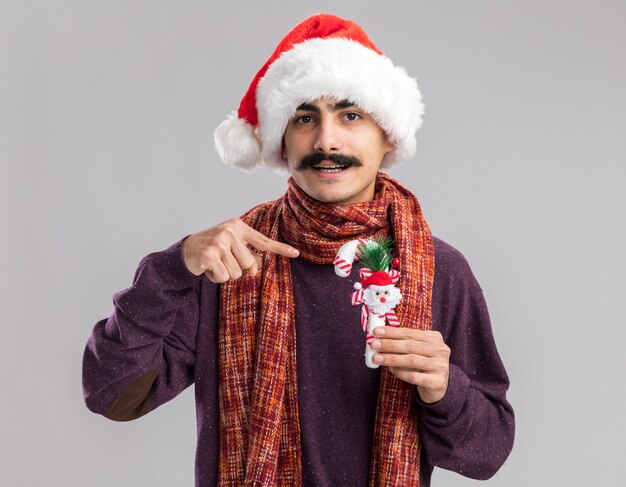 Glücklicher junger Mann mit Schnurrbart, der Weihnachtsmann-Weihnachtsmütze mit warmem Schal um seinen Hals trägt, der Weihnachts-Zuckerstange hält, die mit Zeigefinger lächelnd darauf zeigt