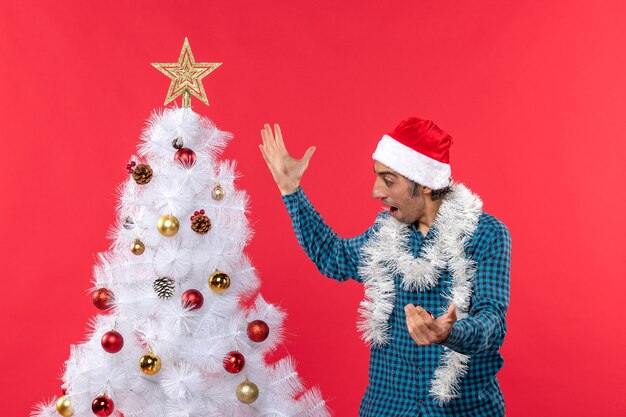 Glücklicher junger Mann mit Weihnachtsmannhut in einem blauen gestreiften Hemd und Blick auf Weihnachtsbaum überraschend rot