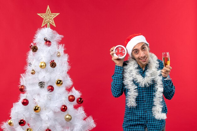 Glücklicher junger Mann mit Weihnachtsmannhut und hält ein Glas Wein und Uhr, die nahe Weihnachtsbaum auf Rot stehen