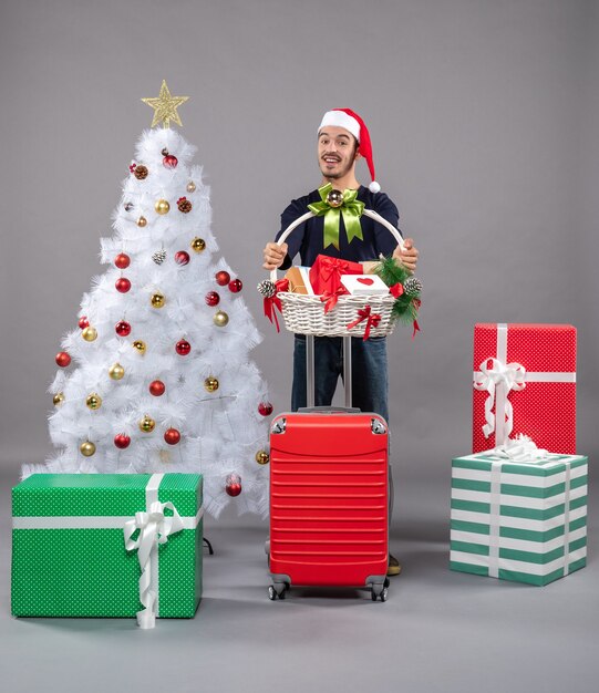glücklicher junger Mann mit Weihnachtsmütze, der Geschenkkorb mit beiden Händen nahe weißem Weihnachtsbaum auf grau hält