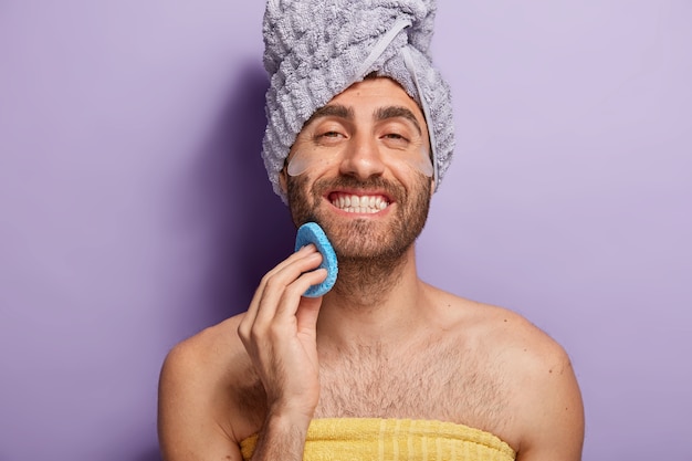 Glücklicher junger Mann pflegt sich nach dem Duschen, will saubere Haut haben, hält Schönheitsschwamm nahe Gesicht, in Handtuch gewickelt, schaut mit Lächeln