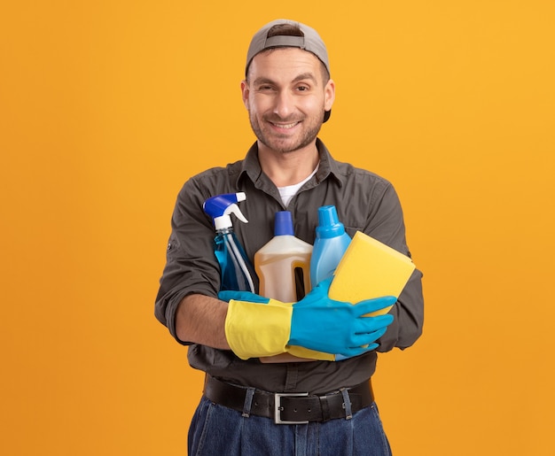 Glücklicher junger reinigungsmann, der freizeitkleidung und kappe in gummihandschuhen hält, die sprühflasche und schwamm halten, die fröhlich über orange wand stehend lächeln