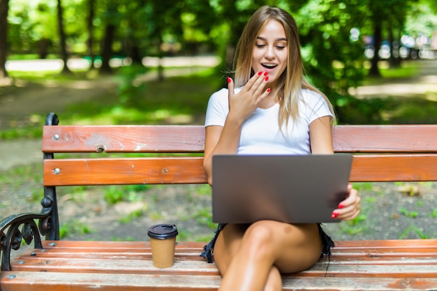 Glücklicher junger Student mit einer Tafel, die auf der Bank sitzt und in einem Sommerpark liest.