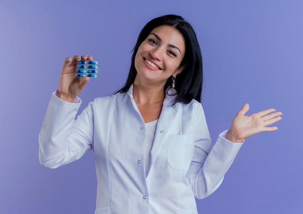 Glücklicher junger weiblicher Arzt, der medizinische Robe trägt Packung der medizinischen Kapseln, die leere Hand suchen und zeigen