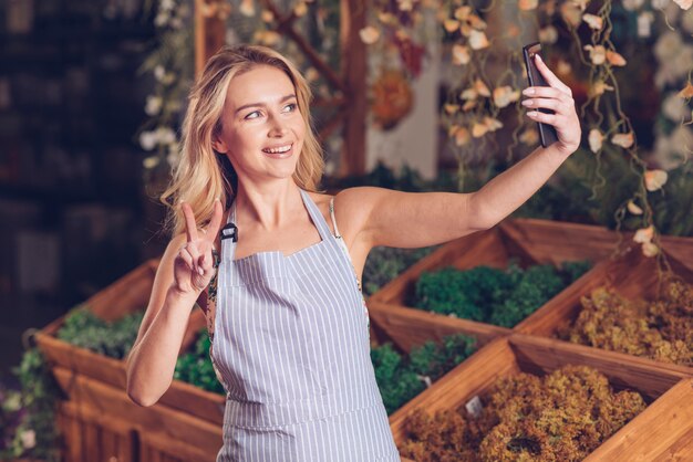 Glücklicher junger weiblicher Florist, der selfie am intelligenten Telefon zeigt Friedensgeste im Shop nimmt