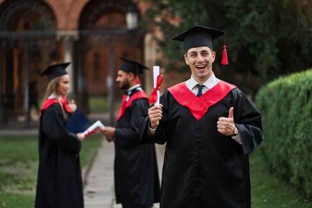 Glücklicher kaukasischer Absolvent mit seinen Klassenkameraden im Abschlusskleid hält ein Diplom auf dem Campus.