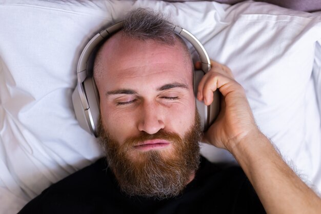 Glücklicher kaukasischer Mann auf dem Bett, der Kopfhörer trägt, hört, genießt seine Lieblingsmusik, ruht sich allein aus, tanzt.