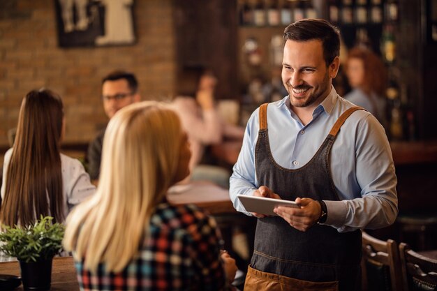 Glücklicher Kellner, der mit weiblichen Kunden spricht und ihre Bestellung auf einem digitalen Tablet in einer Bar schreibt