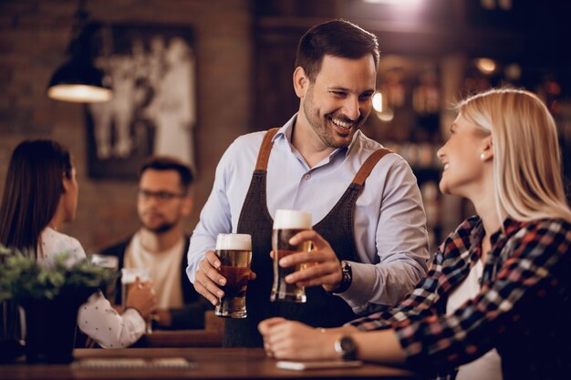 Glücklicher Kellner, der Spaß hat, während er Bier serviert und mit einer Kundin in einer Bar spricht