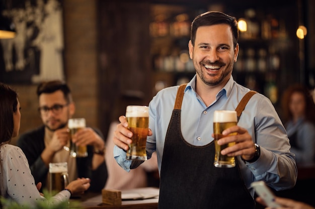 Glücklicher Kellner mit zwei Gläsern Bier und Blick in die Kamera, während er in einer Kneipe arbeitet