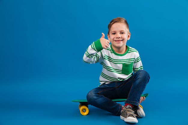 Glücklicher kleiner Junge, der auf Skateboard sitzt und Daumen oben zeigt