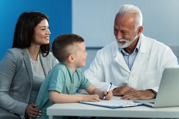 Glücklicher kleiner Junge und seine Mutter kommunizieren während eines Termins in der Arztpraxis mit einem reifen Kinderarzt