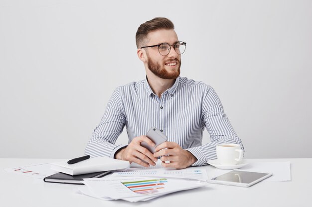 Glücklicher lächelnder bärtiger junger Geschäftsmann schaut zur Seite als Notizkollege, sitzt am Arbeitsplatz mit modernem Smartphone