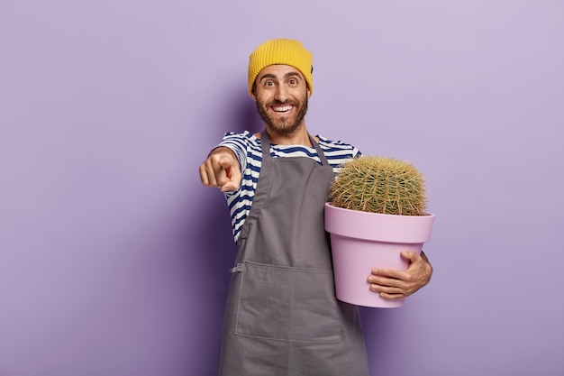 Glücklicher männlicher Gärtner hält Kaktus-Pant Pot, zeigt etwas Fantastisches in der Ferne, zeigt mit dem Zeigefinger, gekleidet in eine spezielle Uniform