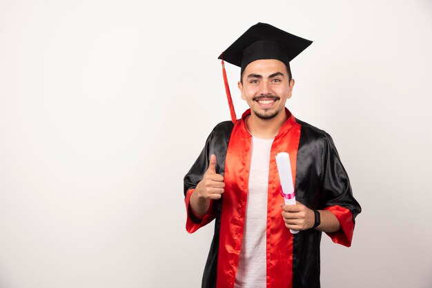 Glücklicher männlicher Student mit Diplom, das Daumen auf Weiß macht.