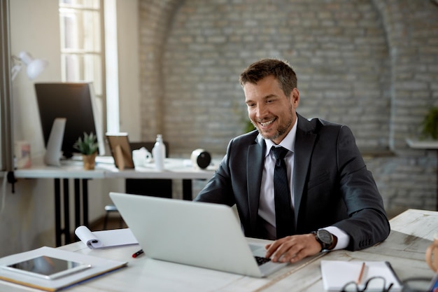 Glücklicher männlicher Unternehmer, der eine E-Mail auf dem Laptop liest, während er im Büro arbeitet